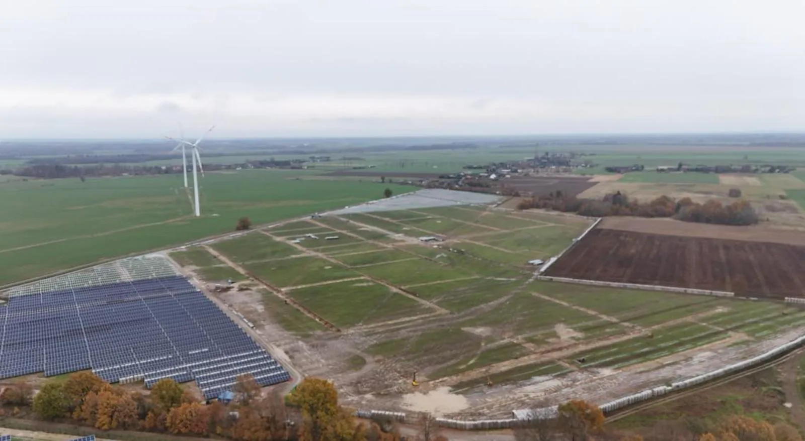 Widok z drona na gotowe pole z modułami fotowoltaicznymi obok terenu budowy kolejnych pól, w tle turbiny wiatrowe