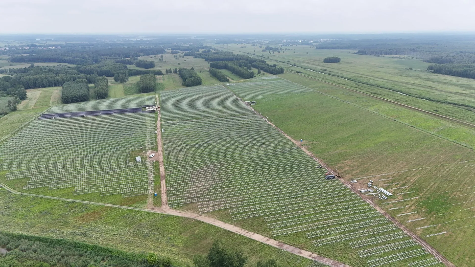 Panorama z drona budowy farmy fotowoltaicznej z rzędami białych stalowych konstrukcji i fragmentem zamontowanych modułów