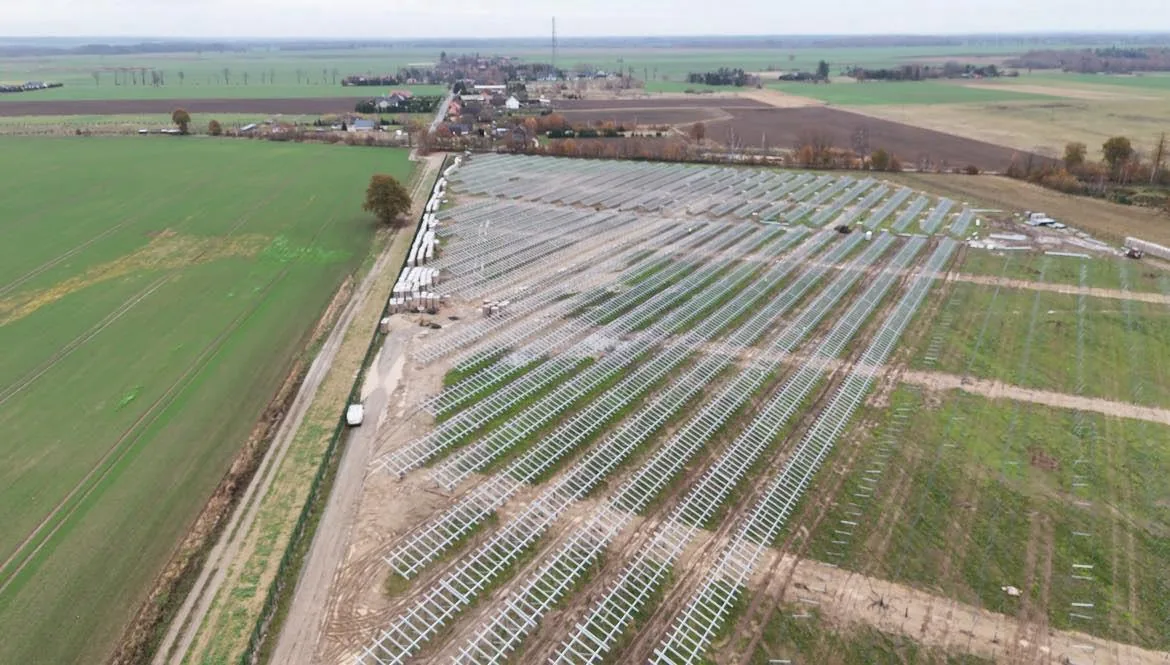 Widok z drona na rzędy stalowych konstrukcji pod moduły fotowoltaiczne na budowie farmy PV, w tle wieś i pola
