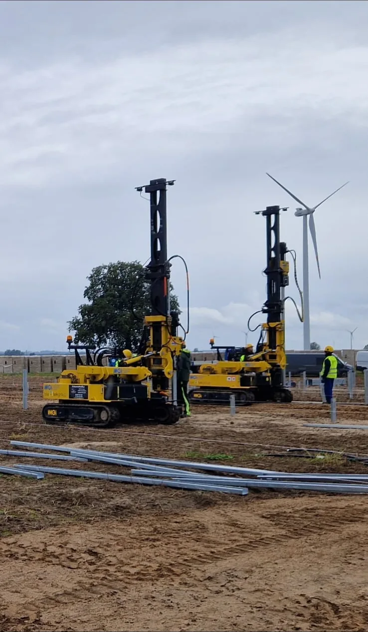 Dwie żółte maszyny do kafarowania na polu budowy, obok pracownicy w odzieży odblaskowej i turbina wiatrowa w tle