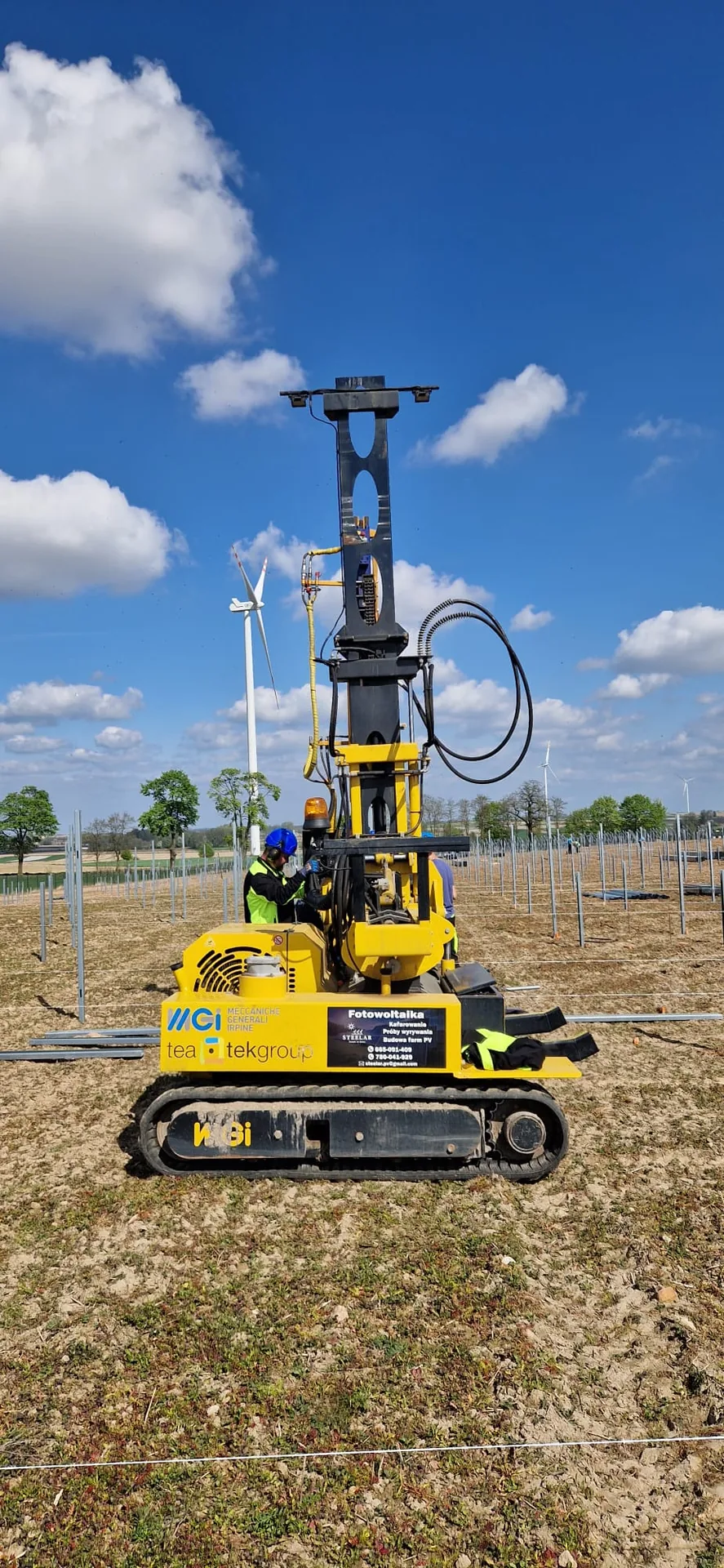 Żółta maszyna do kafarowania z tabliczką z logo STEELAR i listą usług, operator przy maszynie, w tle turbina wiatrowa