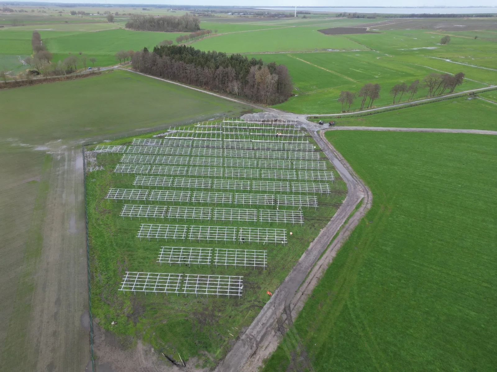 Widok z drona na uporządkowane rzędy białych stalowych konstrukcji pod moduły PV na zielonym polu